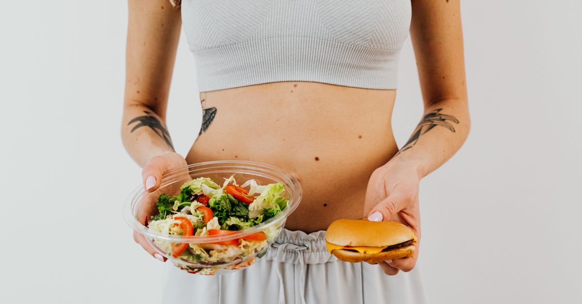 A woman in a sports bra holding a salad and cheeseburger, representing healthy vs junk food choices.